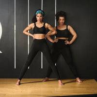 two women in black athletic wear posing against a black wall with white stripes.