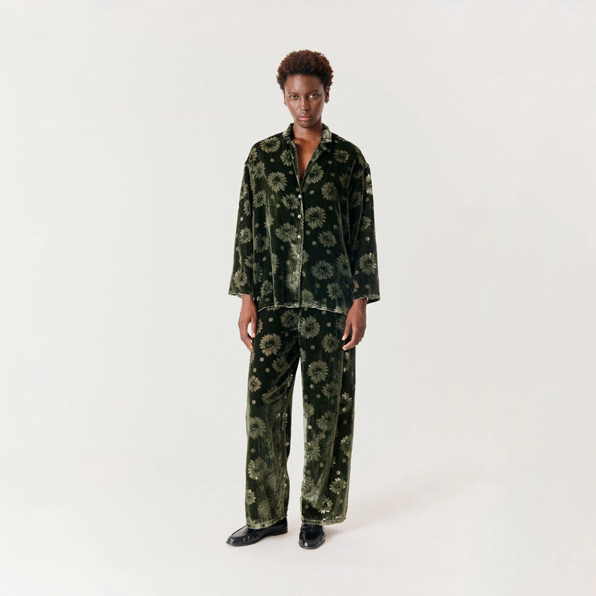 A model stands against a plain light gray studio background wearing a matching two-piece set in dark green velvet with a floral pattern. The set includes a long-sleeve button-up shirt and matching wide-leg pants.