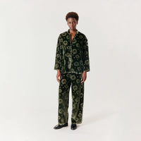A model stands against a plain light gray studio background wearing a matching two-piece set in dark green velvet with a floral pattern. The set includes a long-sleeve button-up shirt and matching wide-leg pants.
