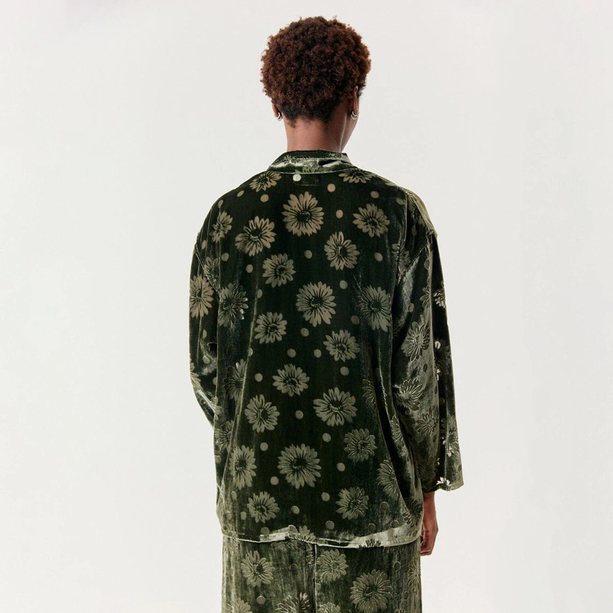 A person viewed from the back wearing a dark green velvet long-sleeve button-up shirt and matching pants with a floral and dot pattern, against a plain white studio background.