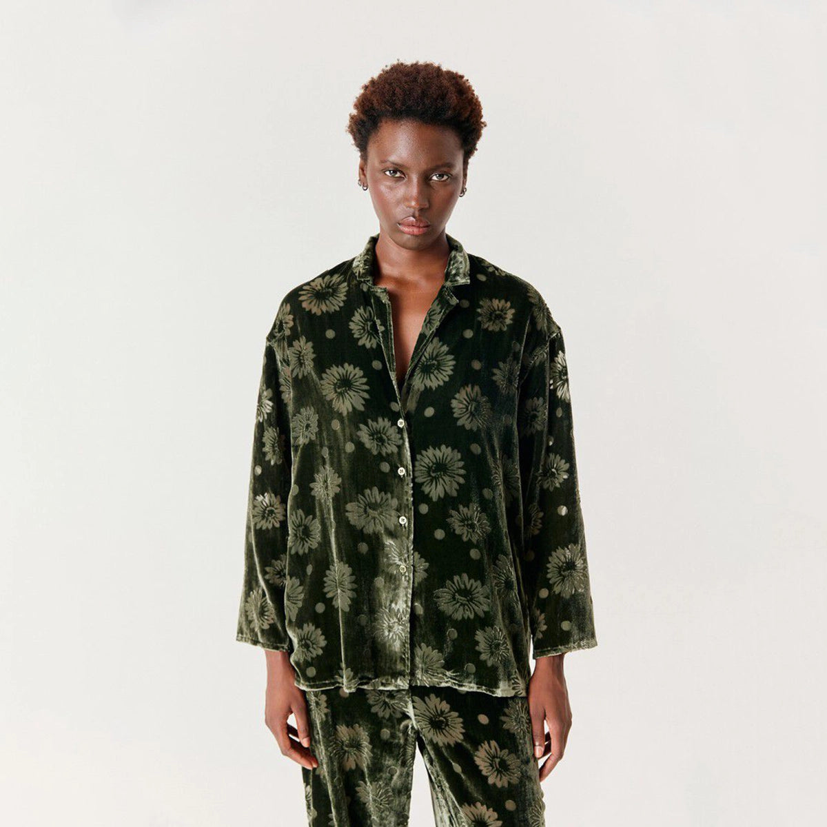 A model wearing a dark green velvet button up shirt with a green floral pattern, consisting of a long-sleeve button-up shirt and matching pants, shot against a plain white studio background.