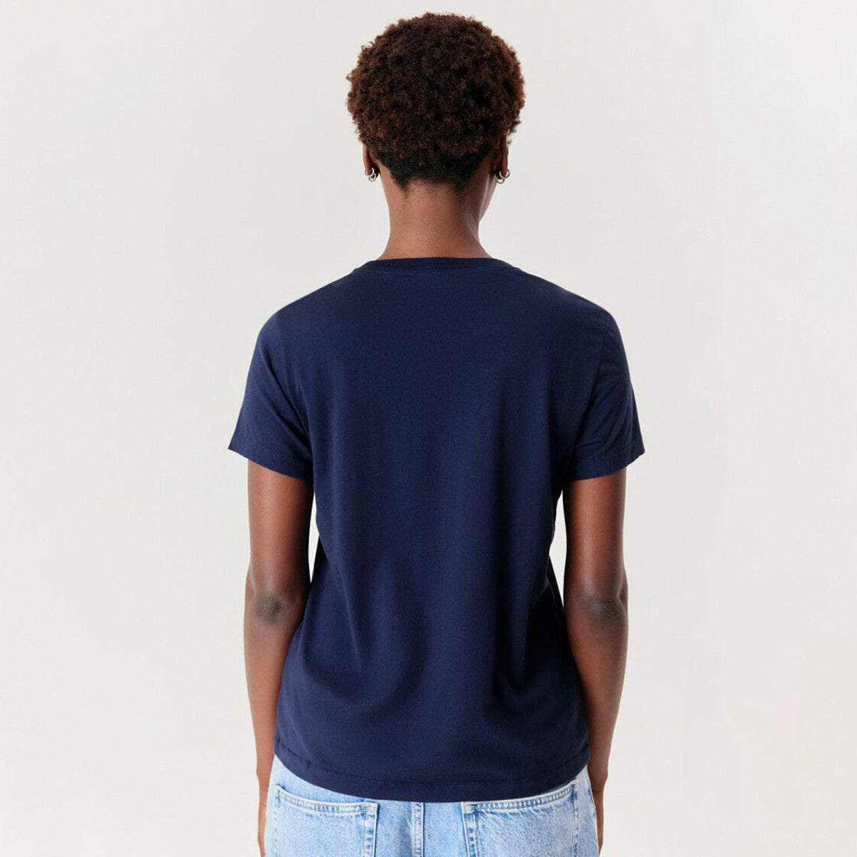 A person shown from the back wearing a plain navy blue short-sleeve crewneck t-shirt and light wash denim jeans, against a clean off-white studio background.