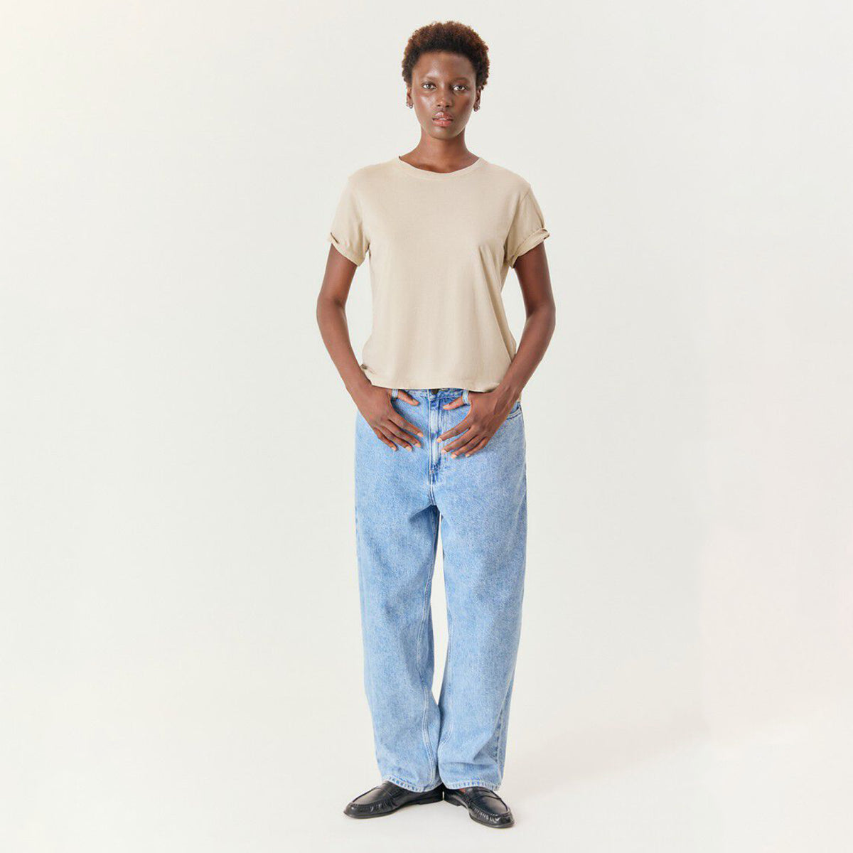 A model stands against a plain off-white studio background wearing a light beige short-sleeve crop top, light blue wide-leg jeans, and black shoes.