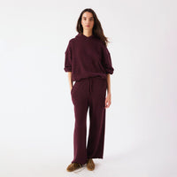 A woman modeling a matching burgundy lounge set consisting of a pullover hoodie and wide-leg sweatpants, paired with brown suede shoes, against a plain white studio background.