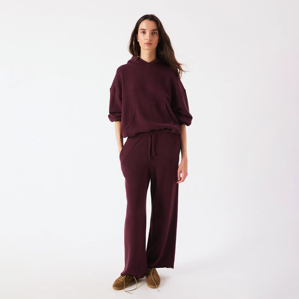 A woman modeling a matching burgundy lounge set consisting of a pullover hoodie and wide-leg sweatpants, paired with brown suede shoes, against a plain white studio background.