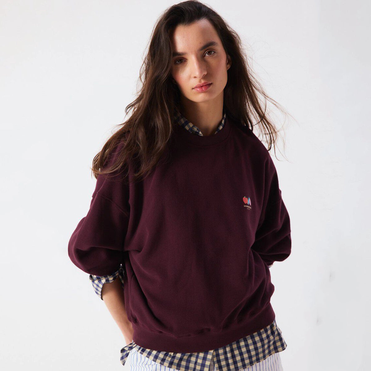 A woman wearing a maroon crewneck sweatshirt with a small embroidered logo on the left chest, layered over a blue and white checkered shirt, against a plain white background.