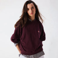 A woman wearing a maroon crewneck sweatshirt with a small embroidered logo on the left chest, layered over a blue and white checkered shirt, against a plain white background.