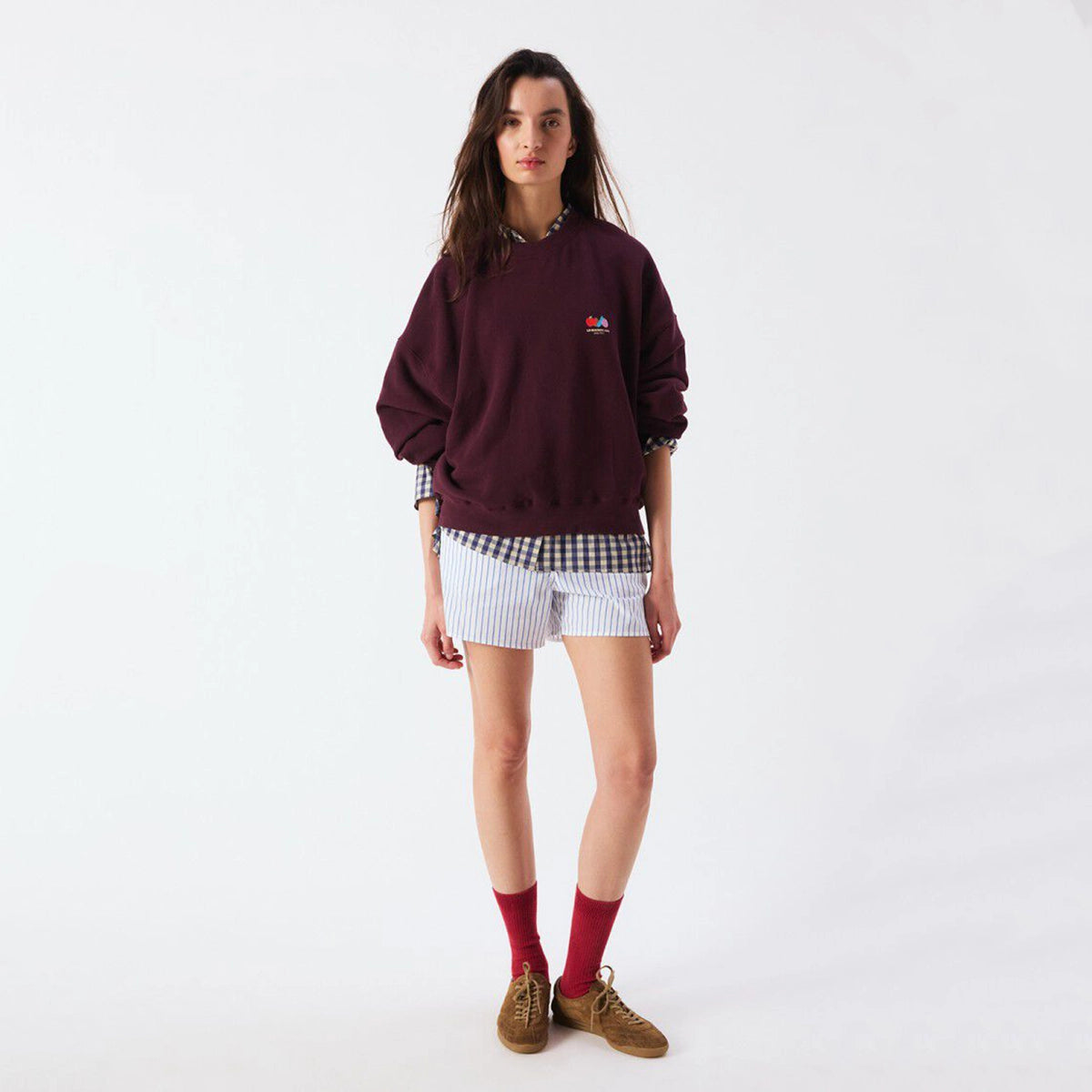 A model stands against a plain white studio background wearing a dark burgundy sweatshirt with a small colorful chest logo, white and navy striped shorts, a black-and-white gingham shirt tucked in, red socks, and brown suede shoes.