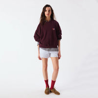 A model stands against a plain white studio background wearing a dark burgundy sweatshirt with a small colorful chest logo, white and navy striped shorts, a black-and-white gingham shirt tucked in, red socks, and brown suede shoes.