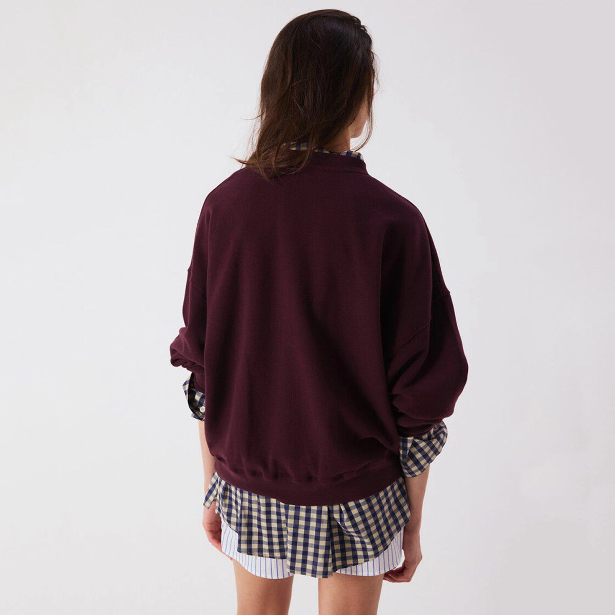 A model shown from the back wearing a dark burgundy crewneck sweatshirt layered over a black and white gingham shirt and striped shorts, against a plain white background.
