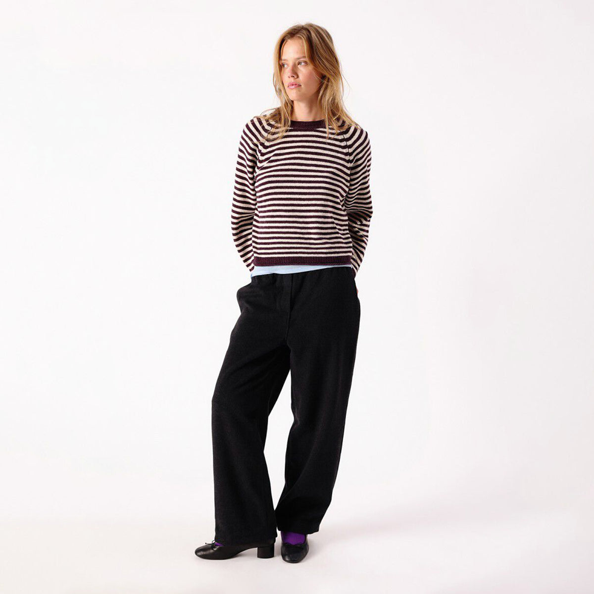 A woman modeling a dark brown and white horizontally striped knit sweater, paired with black wide-leg trousers and black shoes, standing against a plain white studio background.
