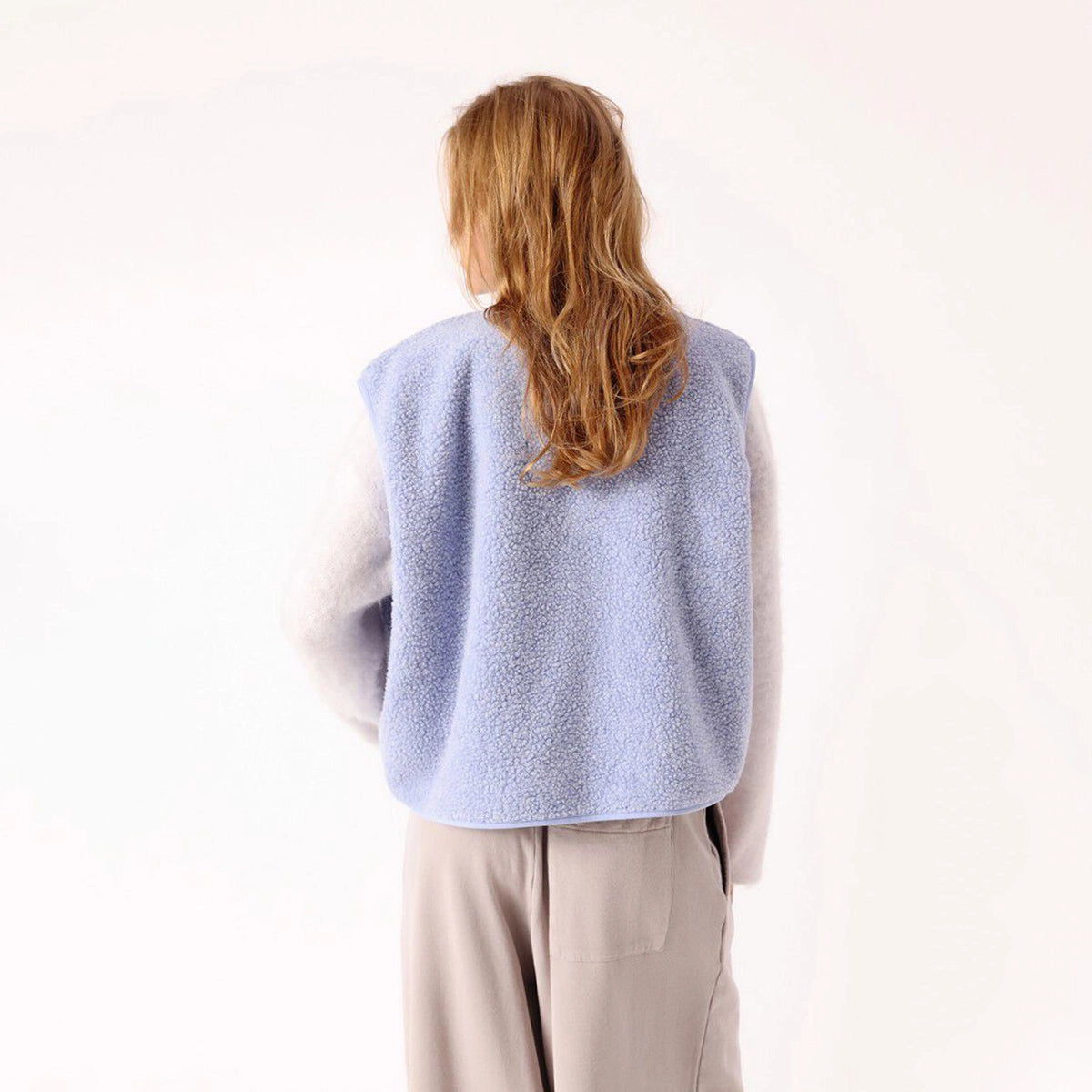 A woman viewed from behind wearing a light blue fuzzy sleeveless vest over a white long-sleeve top and beige trousers, against a plain white background.