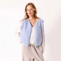 A woman modeling a light blue fleece button-up vest over a white long-sleeve top and beige pants, photographed against a clean white studio background.