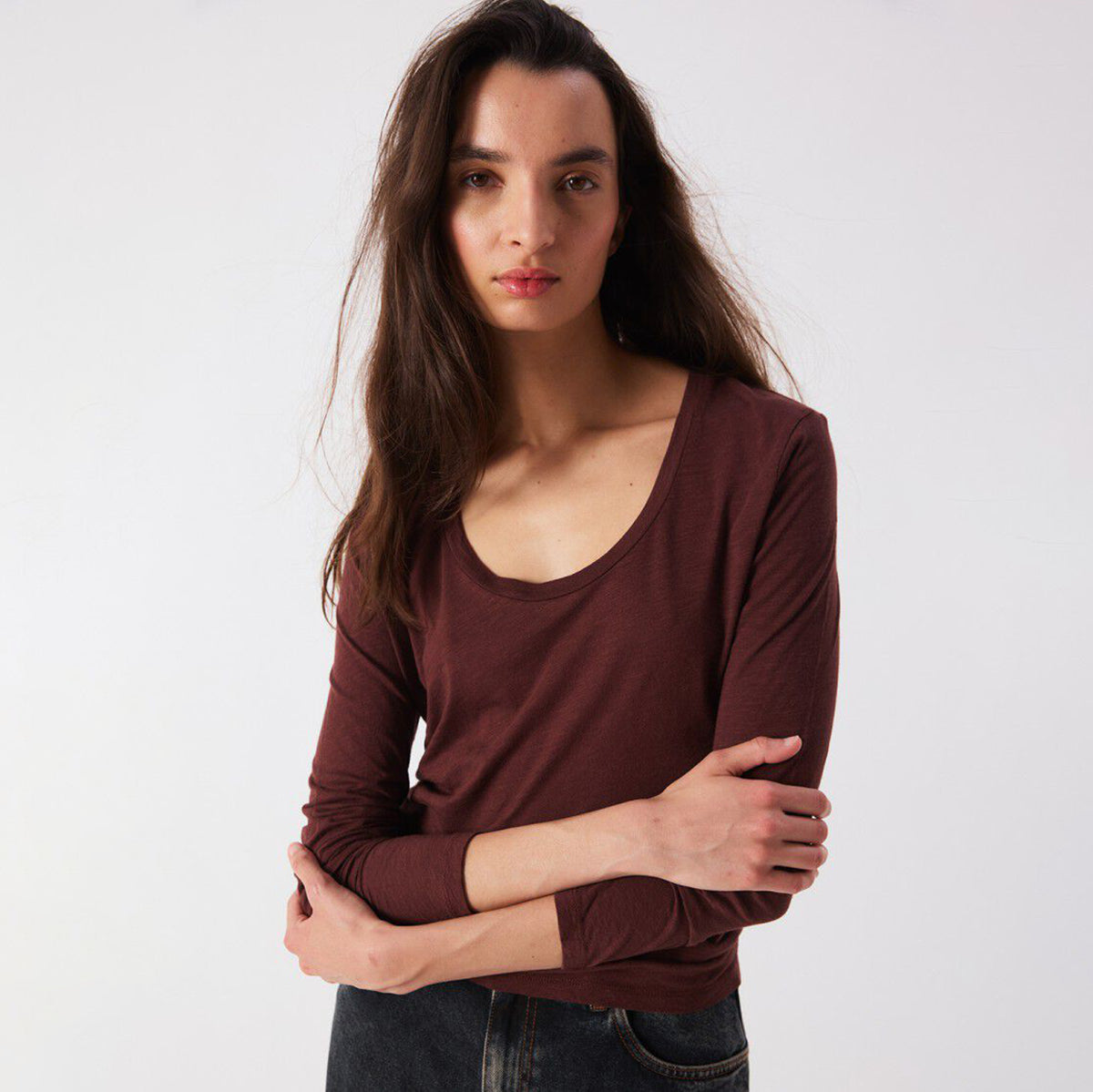 A woman modeling a long-sleeve scoop-neck top in a deep burgundy color, paired with dark jeans, against a plain light gray studio background.