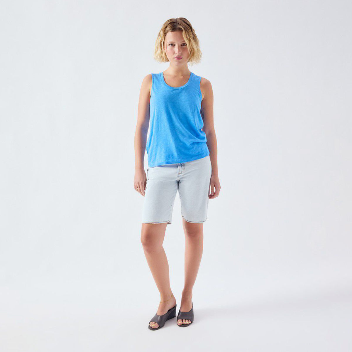 A woman modeling a bright blue sleeveless tank top and light-wash denim shorts against a plain white studio background.