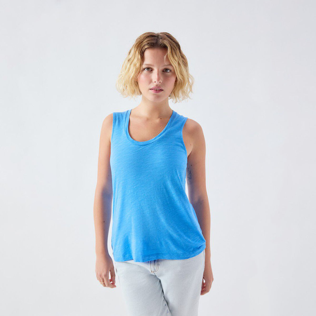 A woman with short blonde hair wearing a bright blue sleeveless scoop-neck tank top and light gray jeans, standing against a plain light gray studio background.