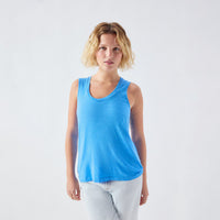 A woman with short blonde hair wearing a bright blue sleeveless scoop-neck tank top and light gray jeans, standing against a plain light gray studio background.