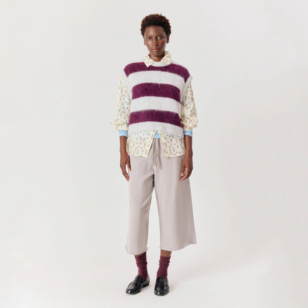 A model stands against a plain off-white studio background wearing a fuzzy striped sweater vest over a cream ruffled shirt with a floral print, paired with light beige cropped trousers, burgundy socks, and black shoes.