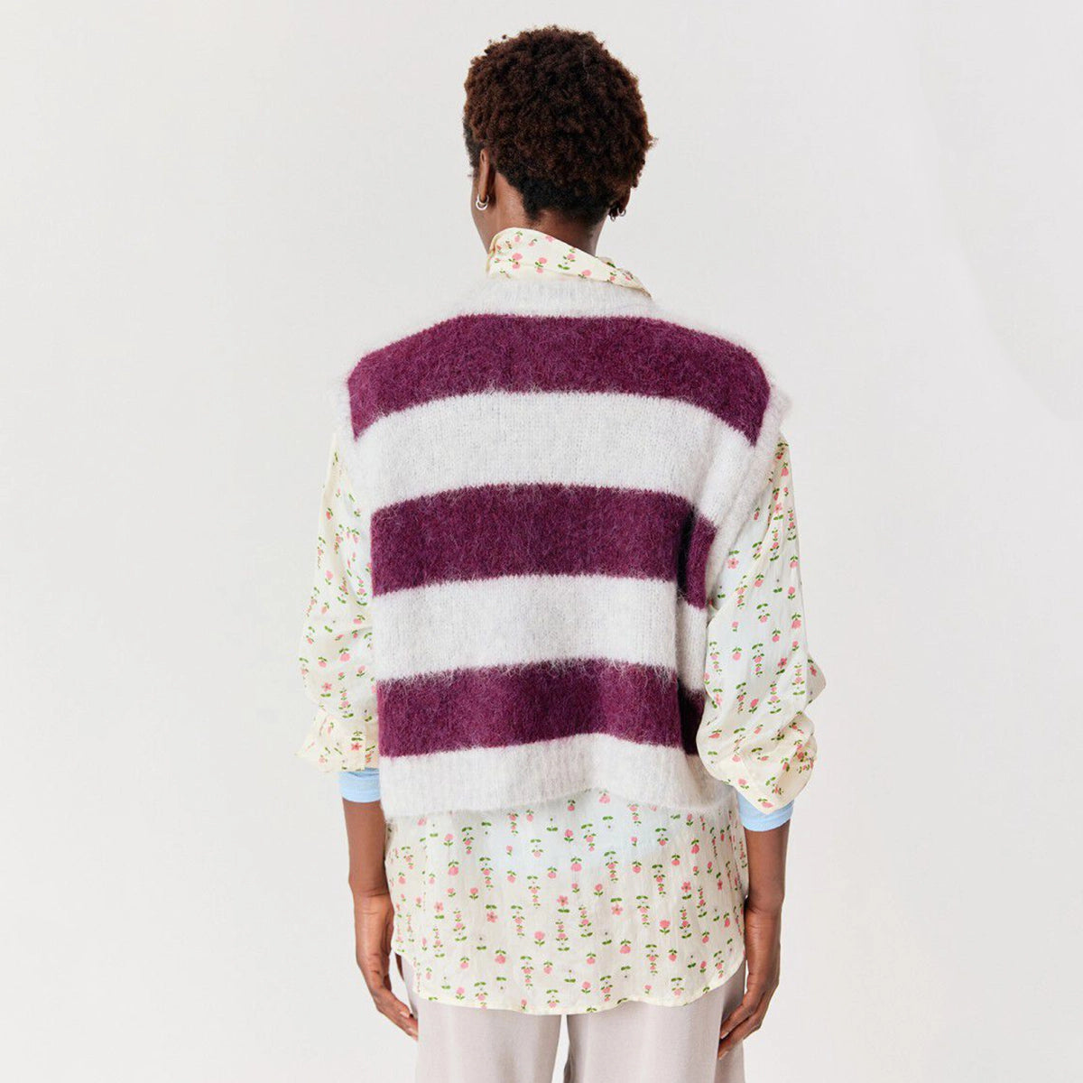 A model shown from the back wearing a fuzzy striped knit vest with horizontal bands of deep purple and off-white, layered over a cream floral shirt.