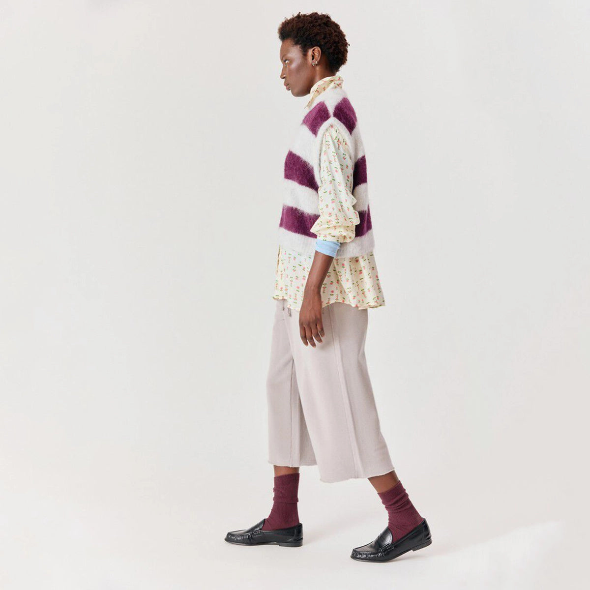 A model is shown from the side in a studio setting, wearing a purple and white striped fuzzy vest over a cream floral shirt, paired with light beige wide-leg trousers, burgundy socks, and black loafers.