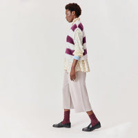 A model is shown from the side in a studio setting, wearing a purple and white striped fuzzy vest over a cream floral shirt, paired with light beige wide-leg trousers, burgundy socks, and black loafers.