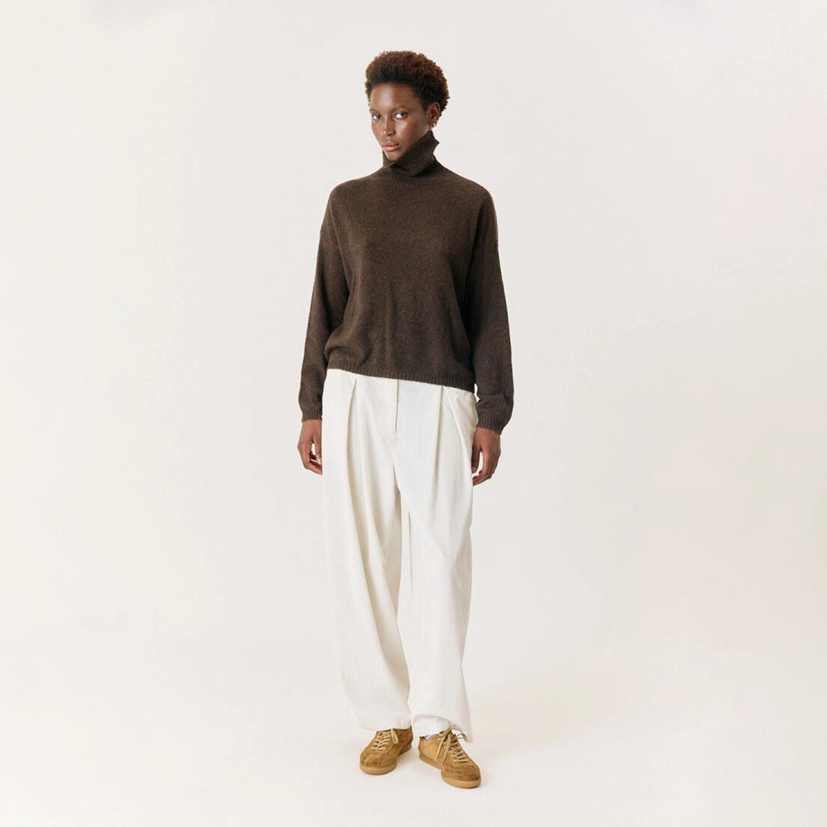 A model stands against a plain off-white studio background wearing a dark brown turtleneck sweater, wide-leg cream trousers, and tan sneakers.