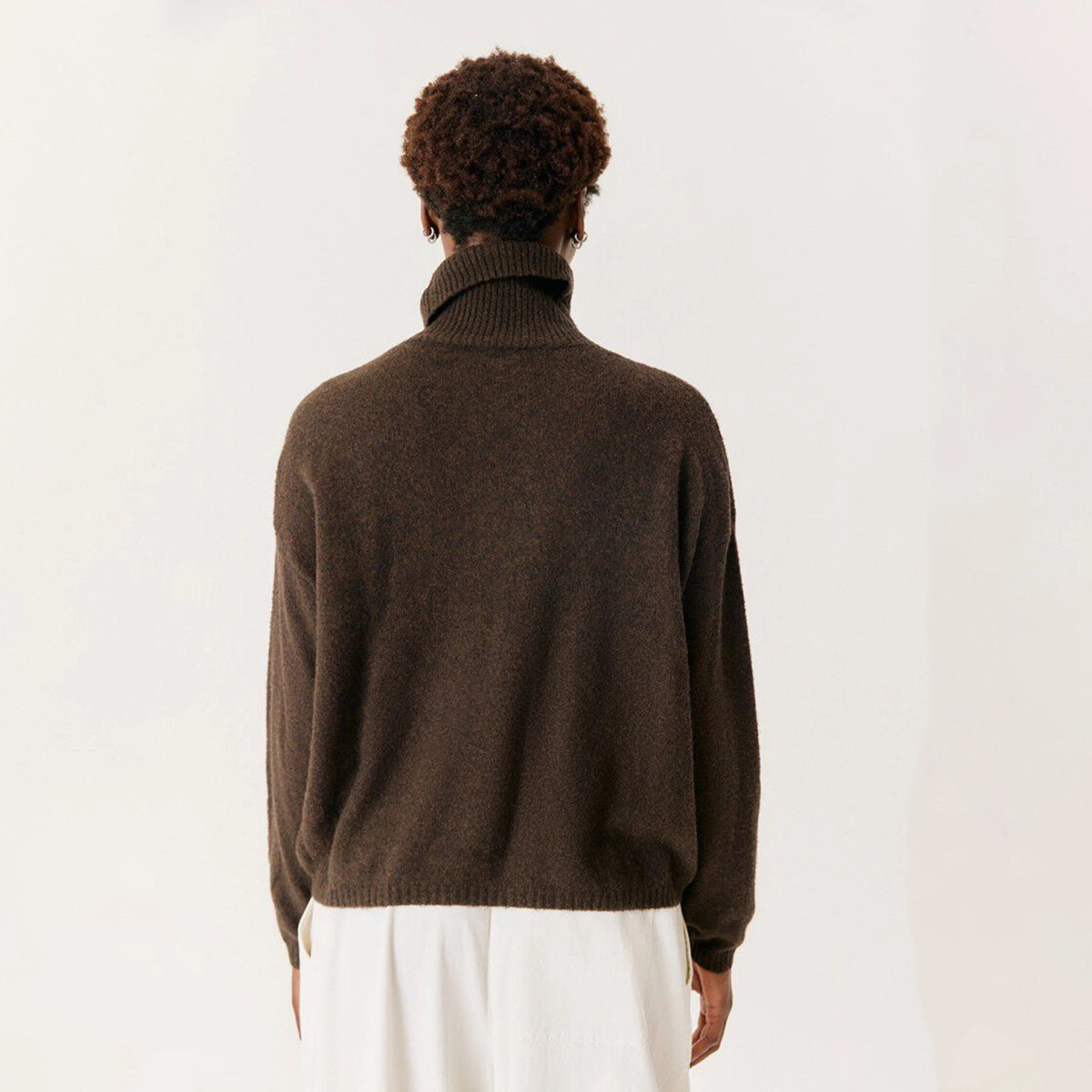 A person viewed from the back wearing a dark brown ribbed turtleneck sweater and white pants against a plain light background.