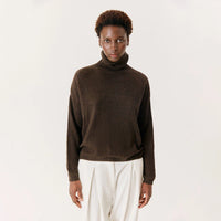 A model wearing a dark brown knit turtleneck sweater and off-white trousers, standing against a plain light gray studio background.