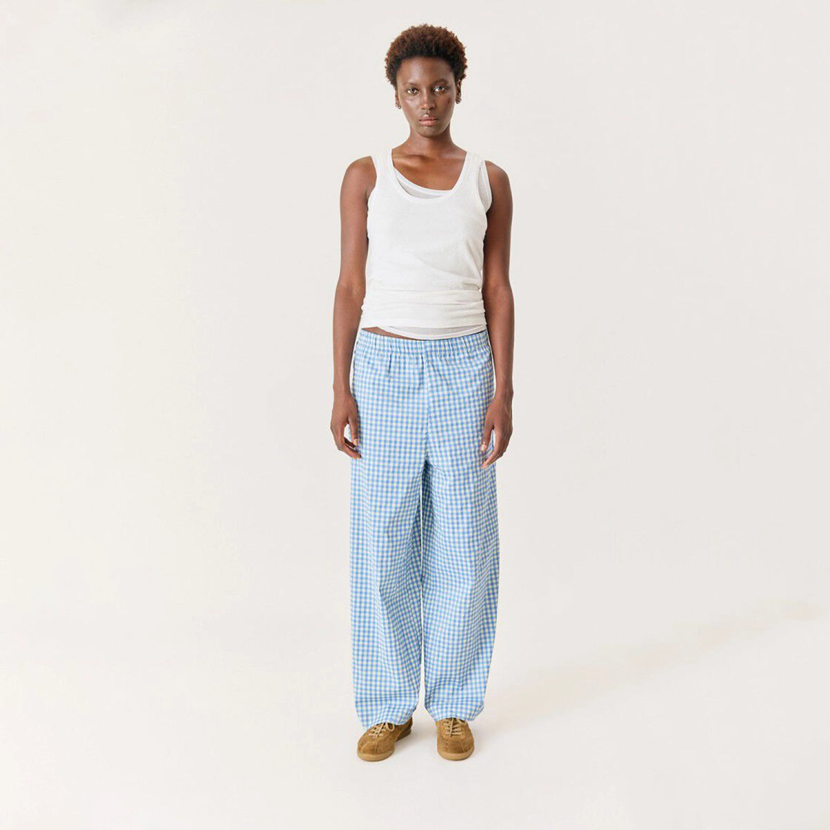 A model stands against a plain off-white studio background wearing light blue and white gingham wide-leg pants, a white tank top, and tan slip-on shoes.