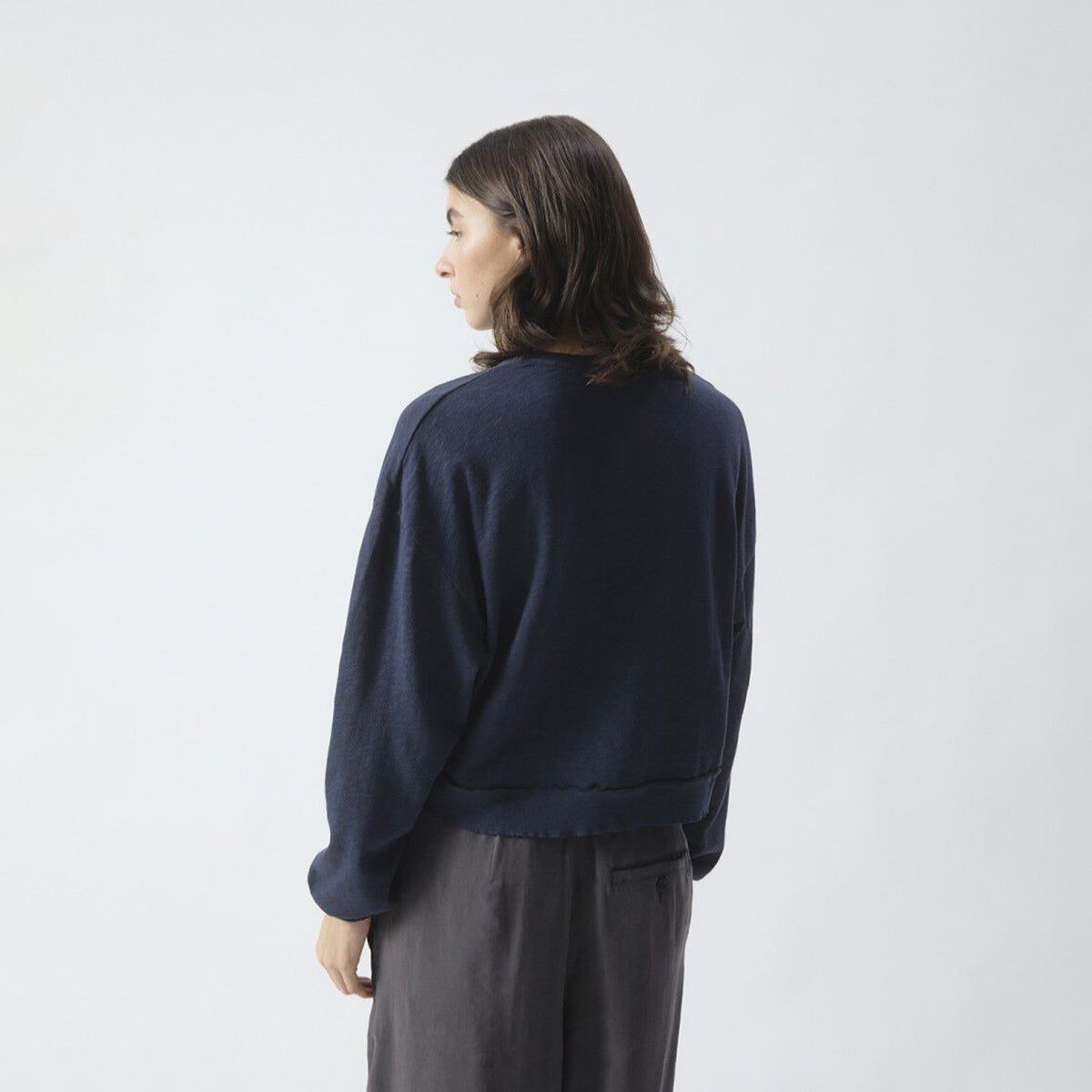 A woman seen from behind wearing a dark navy blue long-sleeve sweatshirt and dark gray pants, standing against a light gray studio background.
