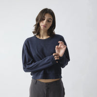 A woman wearing a dark navy blue long-sleeve sweatshirt and dark gray pants, standing against a light gray studio background.