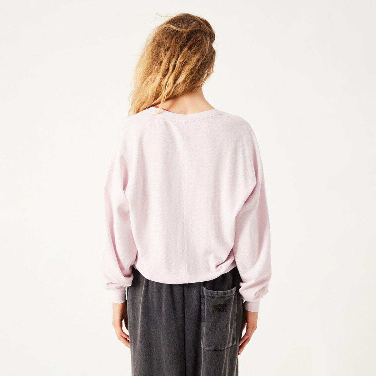 A woman viewed from behind wearing a light pink long-sleeve crewneck sweatshirt and dark gray wide-leg pants against a plain white studio background.