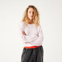 A woman with curly blonde hair stands against a plain white studio background, wearing a light pink crewneck sweatshirt over a bright orange-red undershirt and dark charcoal gray pants.