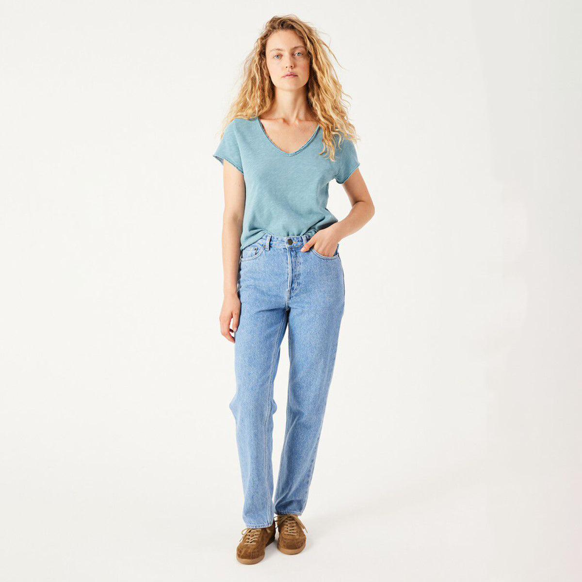 A woman modeling a light blue v-neck t-shirt and light-wash blue jeans with brown suede shoes, standing against a plain white studio background.