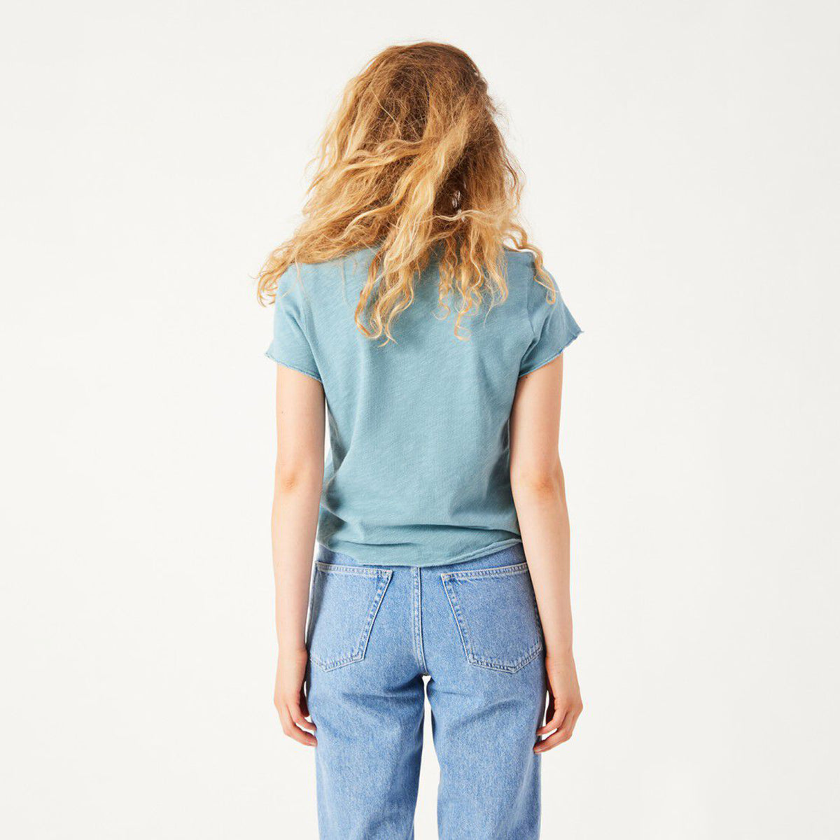 A woman with long, wavy blonde hair is shown from behind wearing a heathered slate blue t-shirt and light wash blue jeans against a plain white studio background.