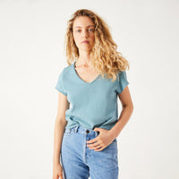 A woman with curly blonde hair wearing a light blue v-neck t-shirt and light wash blue jeans, standing against a plain white studio background.