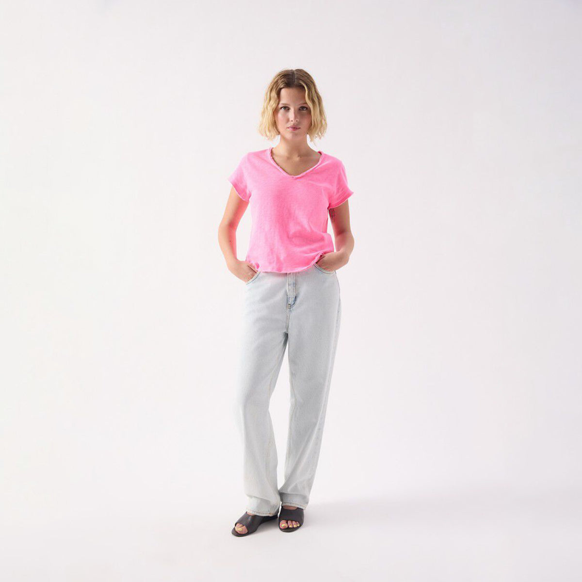 A woman modeling a bright pink short-sleeve V-neck t-shirt and light gray wide-leg jeans against a plain white studio background.