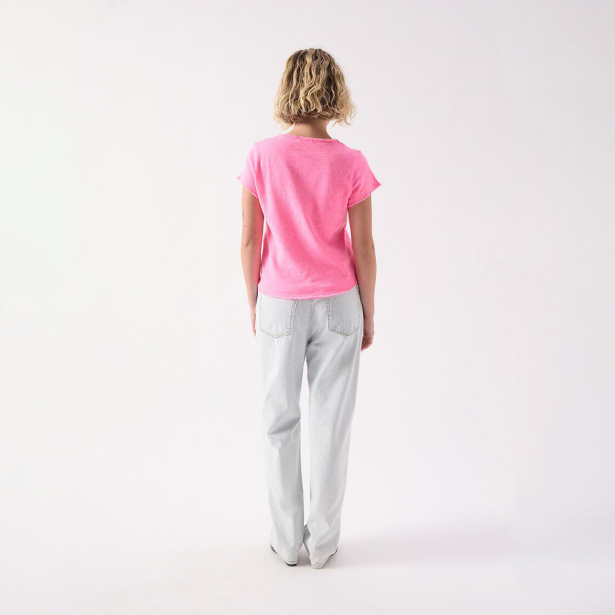 A woman viewed from behind wearing a bright pink short-sleeve t-shirt and light wash jeans, standing against a plain white studio background.
