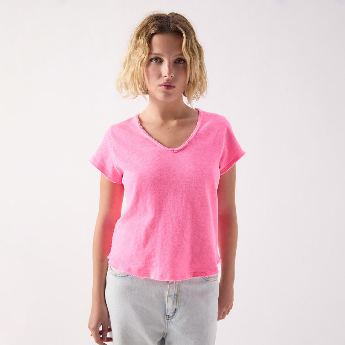 A woman with short blonde hair wearing a bright pink v-neck t-shirt and light-wash jeans, standing against a plain white studio background.