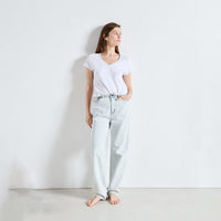 A woman stands barefoot against a plain white wall wearing a white V-neck t-shirt and light-wash wide-leg jeans.