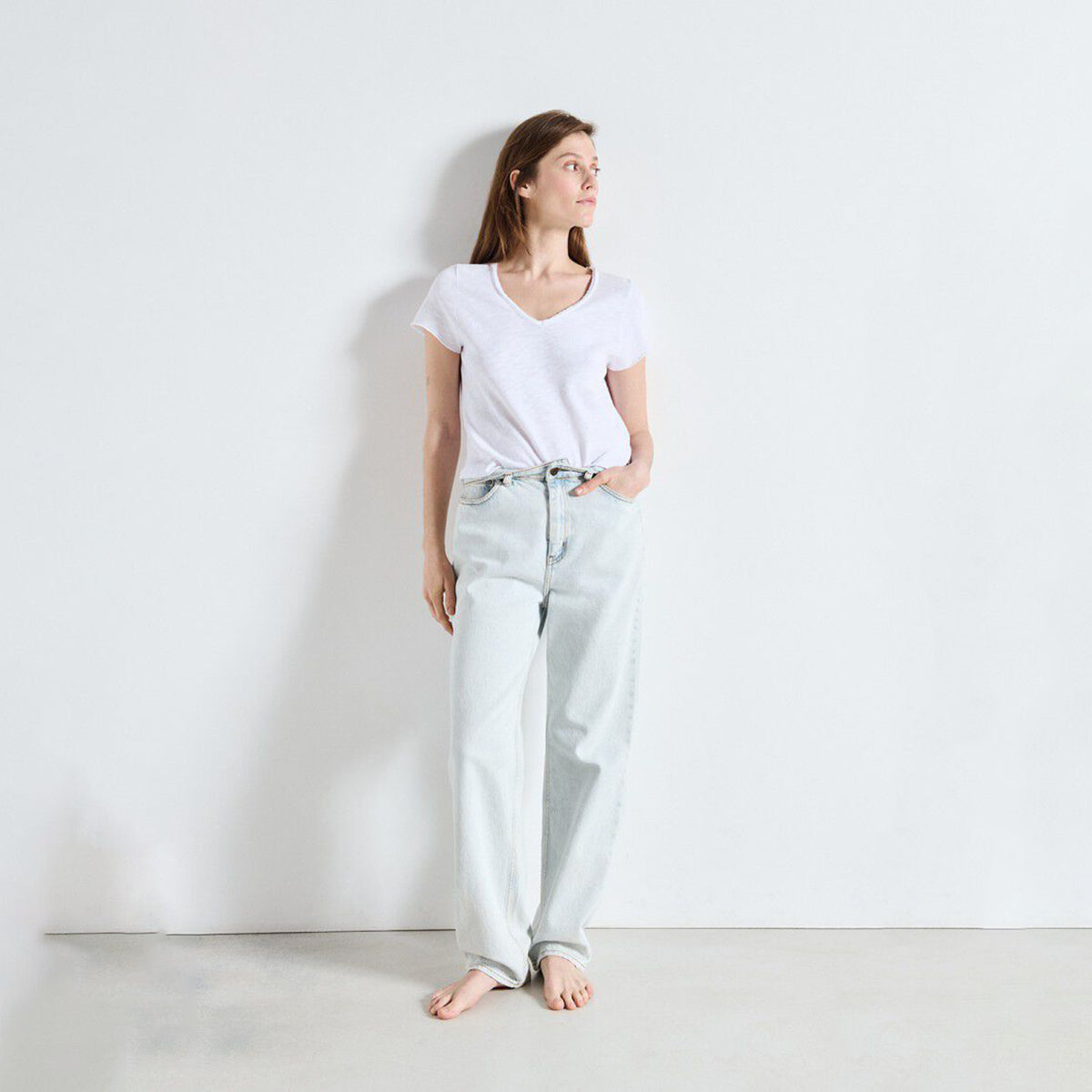A woman stands barefoot against a plain white wall wearing a white V-neck t-shirt and light-wash wide-leg jeans.
