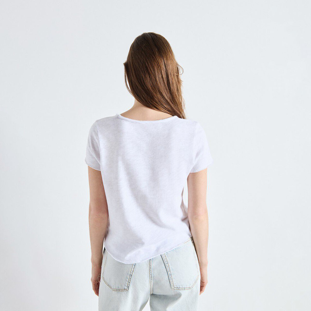 A woman viewed from behind wearing a plain white short-sleeve t-shirt and light-wash jeans against a clean white studio background.