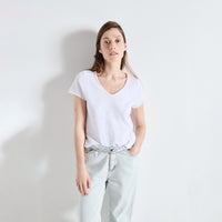 A woman modeling a white short-sleeve V-neck t-shirt tucked into light-wash jeans, standing against a plain white wall.