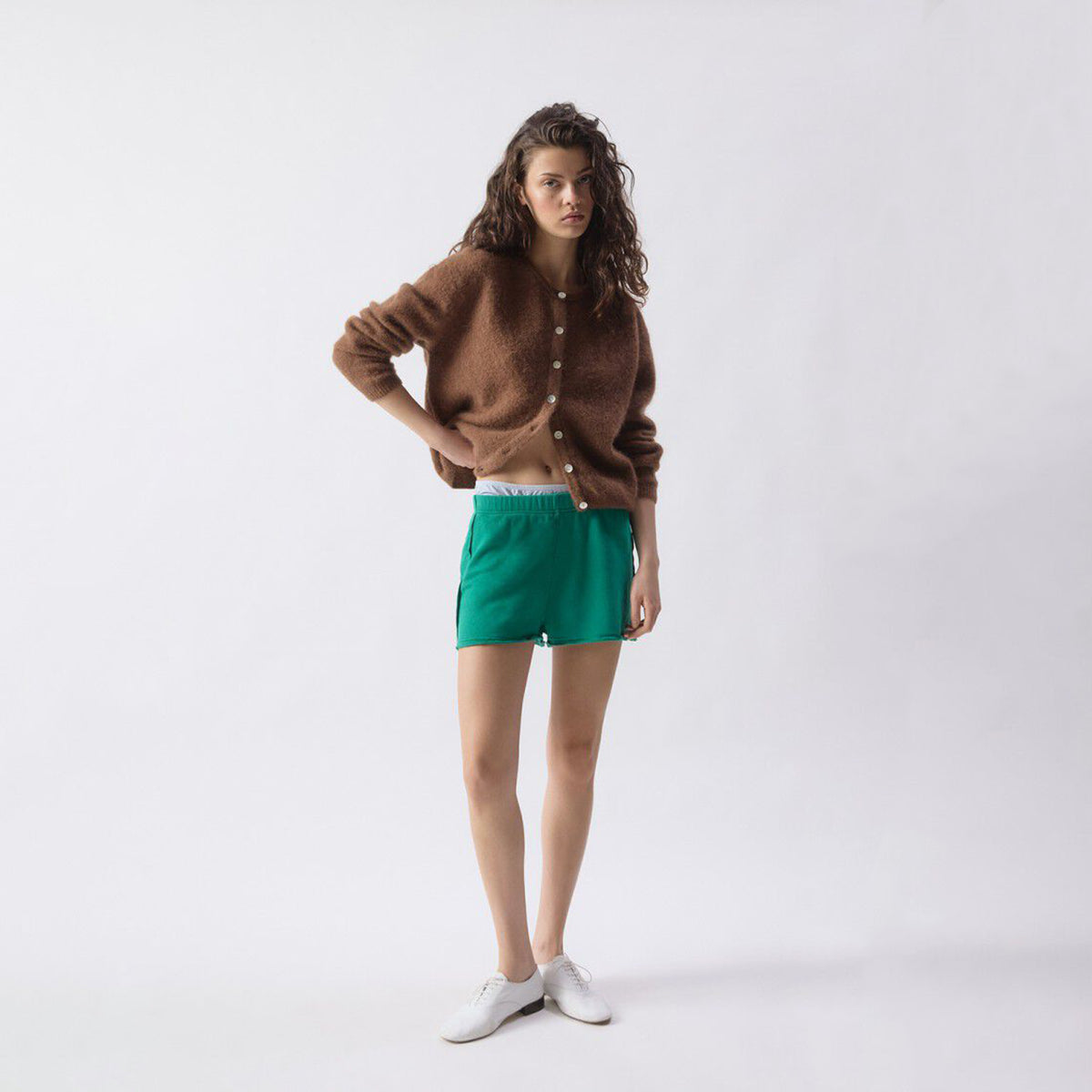 A model stands against a plain light gray studio background wearing a brown fuzzy button-front cardigan, bright green shorts, and white sneakers.