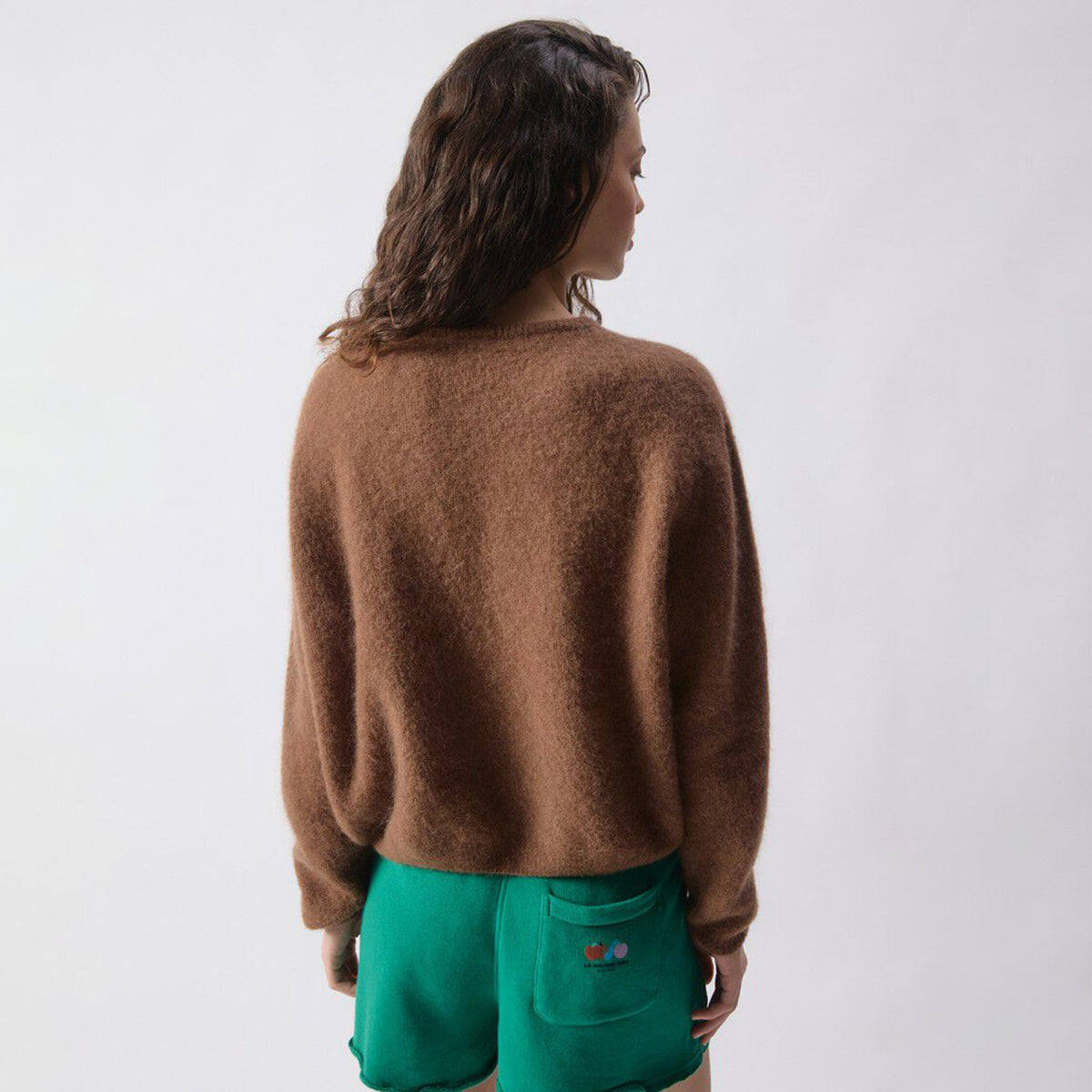 A woman shown from the back wearing a fuzzy brown crewneck cardigan and bright green shorts against a plain light gray studio background.