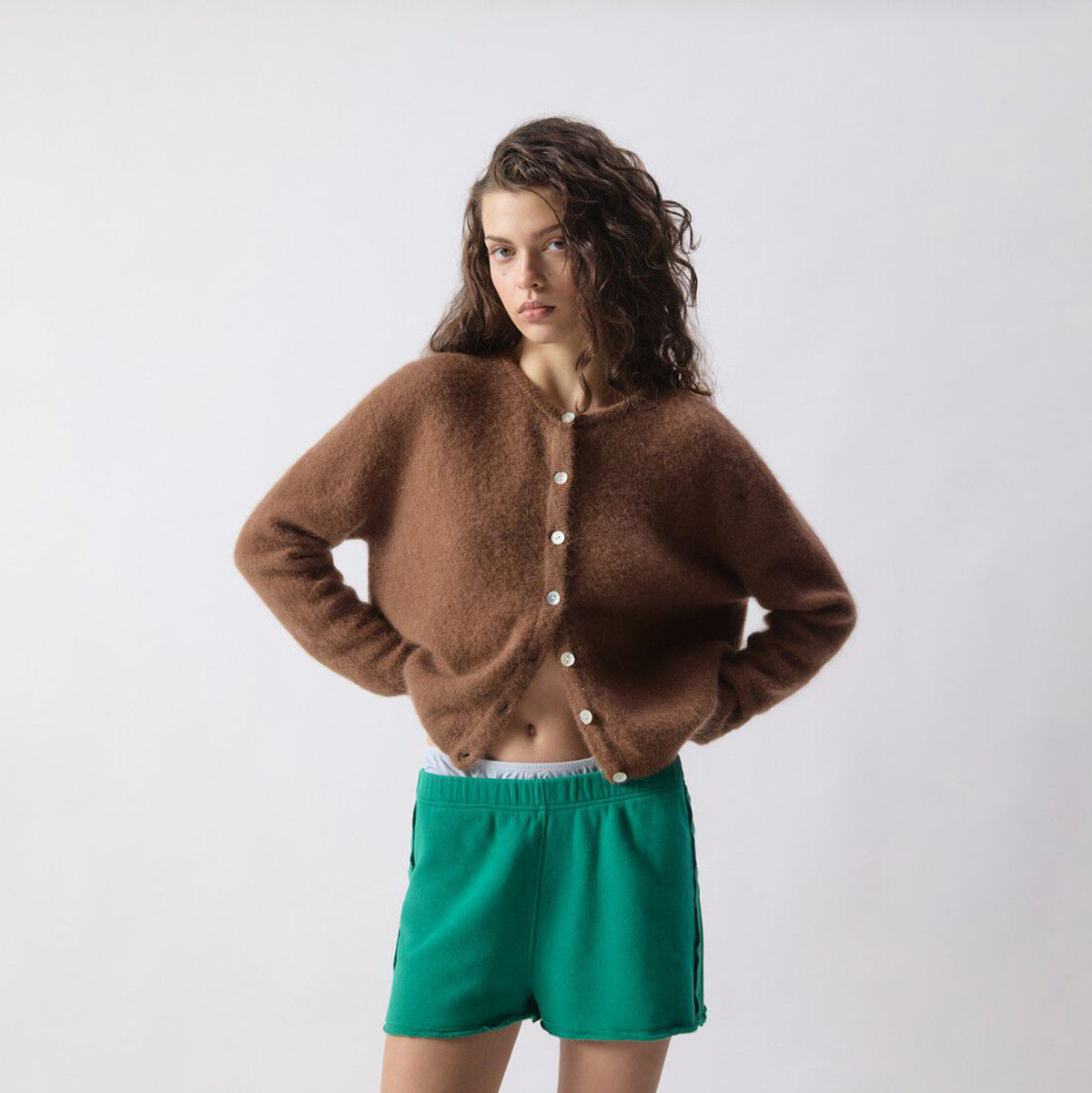 A model wearing a fuzzy brown button-up cardigan and bright green shorts, standing against a plain light gray studio background.