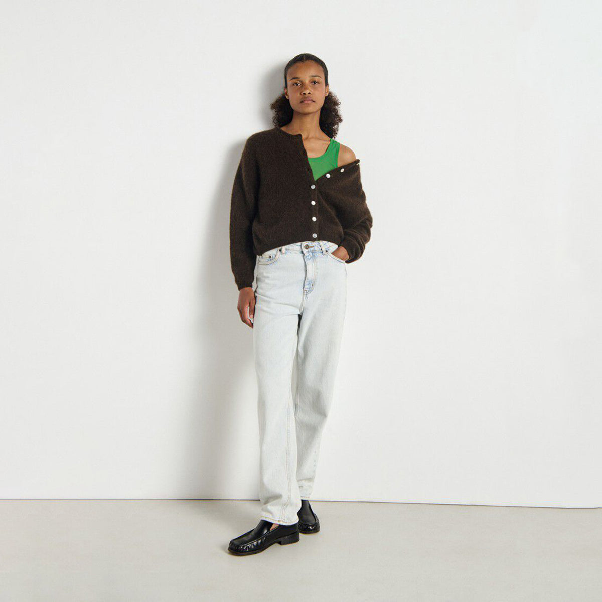 A model stands against a plain white wall wearing a dark brown knit cardigan with asymmetrical button details, light-wash wide-leg jeans, and black loafers.