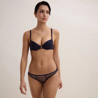 A woman wearing a navy blue lingerie set with gold-tone hardware and sheer patterned mesh, photographed against a plain light beige studio background.