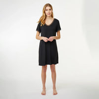 A woman modeling a short-sleeve black nightgown with a V-neck and delicate lace trim, standing against a plain white studio background.