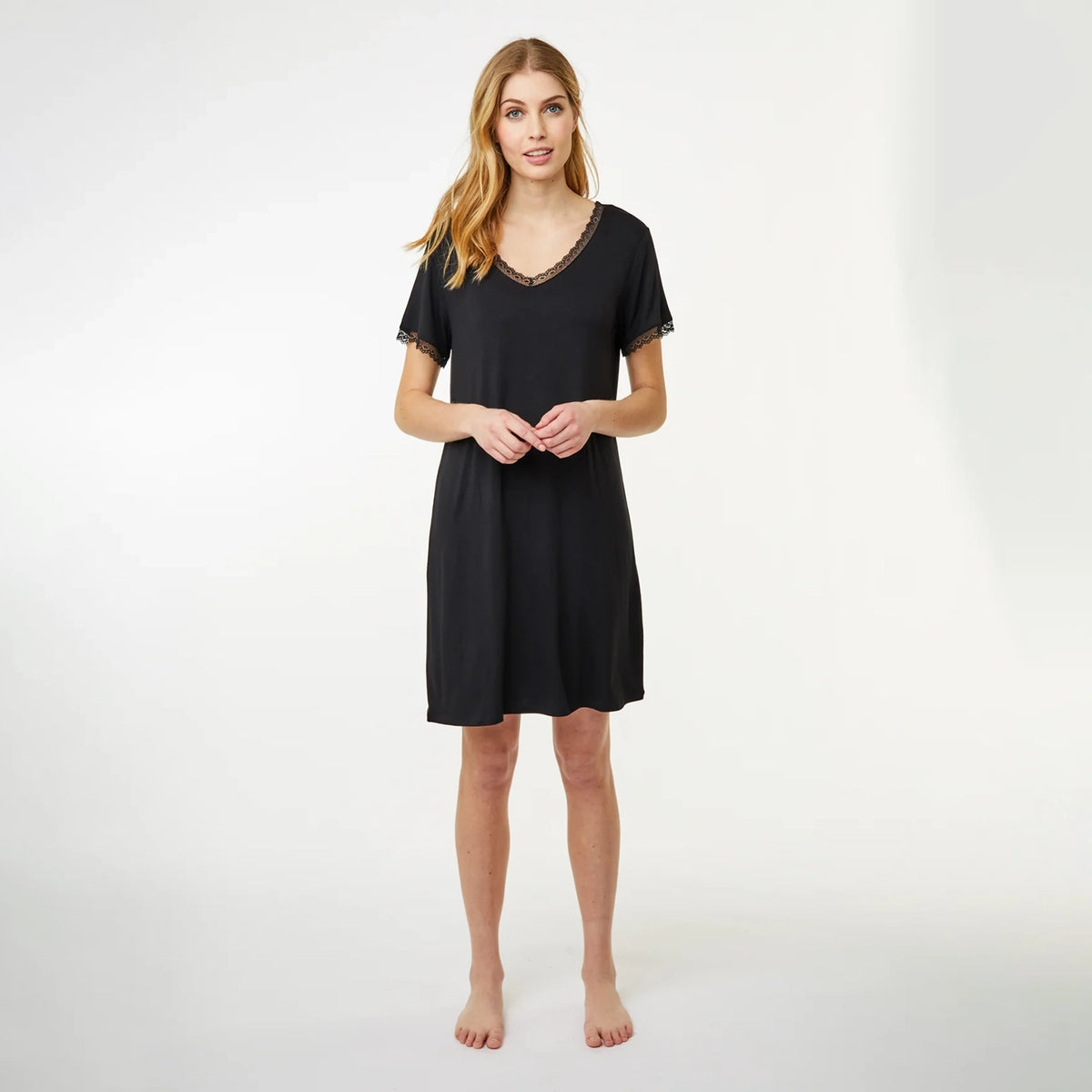 A woman modeling a short-sleeve black nightgown with a V-neck and delicate lace trim, standing against a plain white studio background.